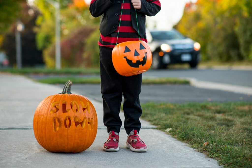 Child in costume holding a jack-o-lantern candy bucket on a sidewalk beside a carved pumpkin reading SLOW DOWN, with a car approaching